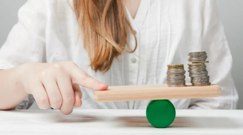 Une femme tient une pile de pièces sur une balance en bois, symbolisant l'équilibre financier et la gestion des dettes.