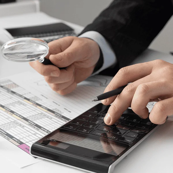 Un homme en costume utilise une calculatrice et une loupe pour examiner des documents.