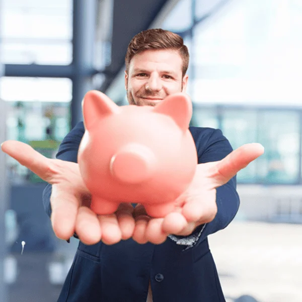 Un homme en costume tient une tirelire en forme de cochon, souriant et regardant vers l'objectif.