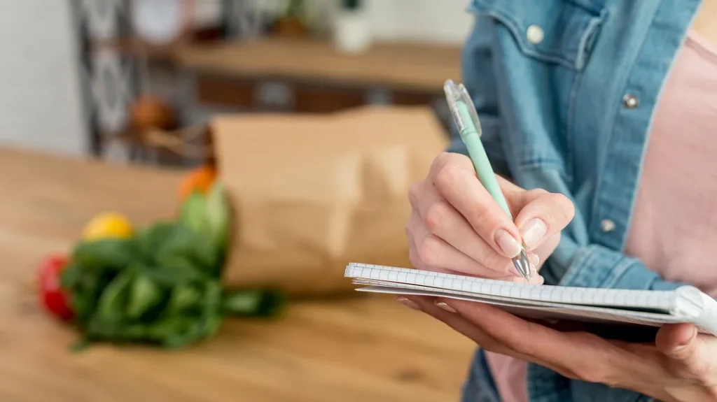 Femme écrivant dans un carnet avec un stylo, des légumes en arrière-plan.
